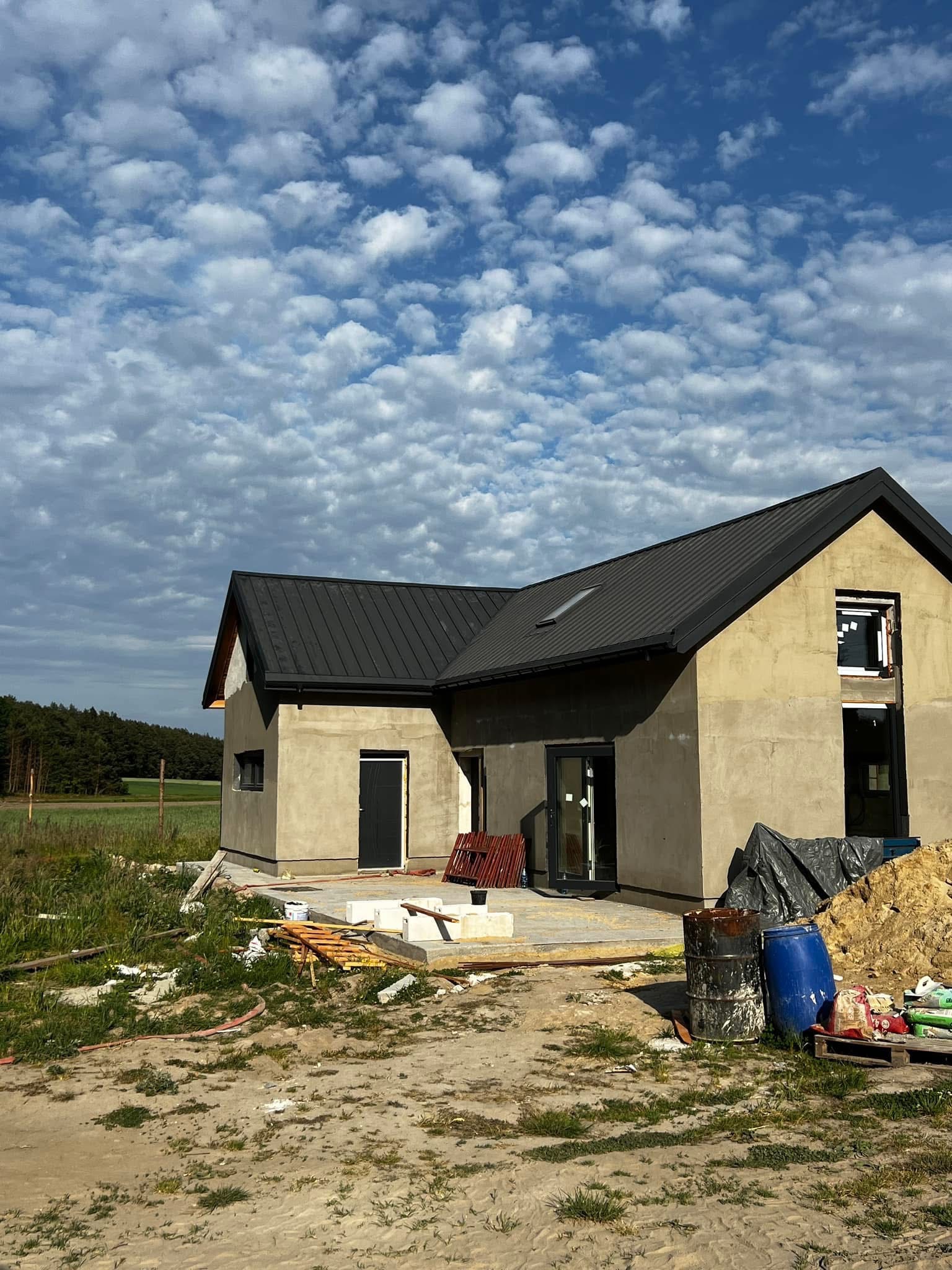 Dom jednorodzinny w trakcie budowy, z czarnym dachem z blachy na tle błękitnego nieba z chmurami. Widoczne materiały budowlane i teren wokół domu.