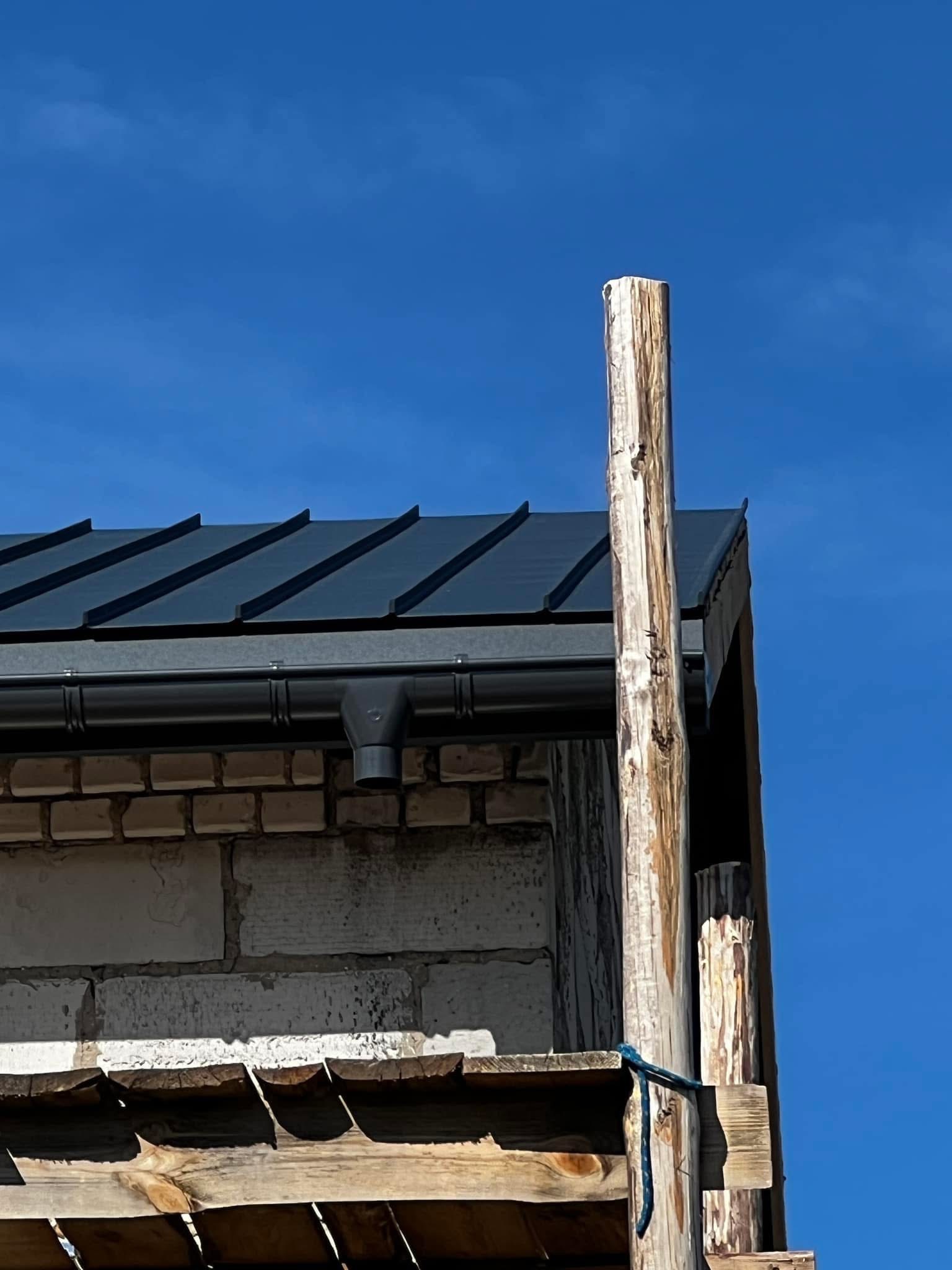 Fragment dachu z blachy w kolorze grafitowym, z zamontowaną rynną i rurą spustową, widoczne z rusztowania na tle błękitnego nieba.