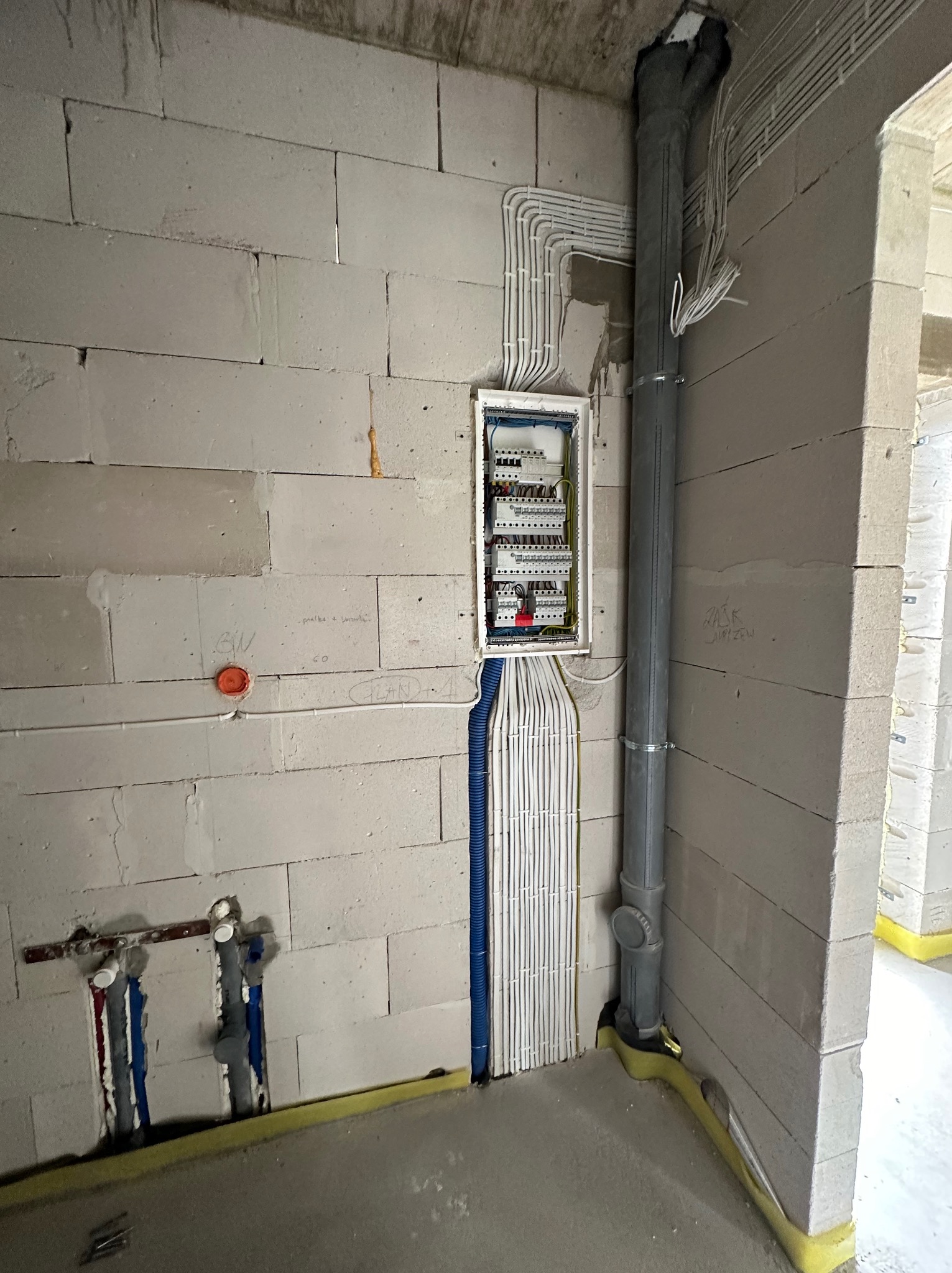 Rozdzielnia elektryczna z poprowadzonymi przewodami w surowym wnętrzu budynku z betonu komórkowego. Widoczne rury kanalizacyjne i instalacje wodne.
