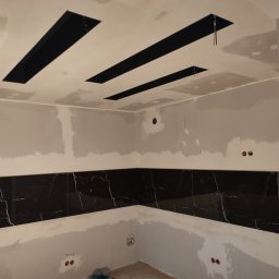 Usługi budowlane - Wnętrze w trakcie prac wykończeniowych: czarne, marmurowe płytki ułożone w pasie na ścianie, sufit z otworami na oświetlenie, ściany przygotowane do malowania, widoczne ślady szpachlowania.