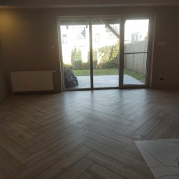 Usługi budowlane - Ułożona podłoga z paneli w jodełkę w nowym pokoju z dużym oknem tarasowym wychodzącym na ogród, widoczny grzejnik na ścianie i odkurzacz przemysłowy.