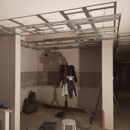 Usługi budowlane - Montaż metalowej konstrukcji sufitu podwieszanego w trakcie prac remontowych, widoczne narzędzia, drabina i ubrania na wieszaku.