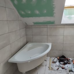 Usługi budowlane - Narożna wanna na nóżkach w trakcie montażu w łazience z szarymi płytkami, sufit z płyt karton gips, widoczne narzędzia i materiały budowlane.