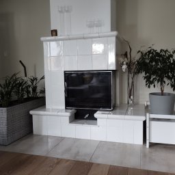 Usługi budowlane - Biały, kaflowy kominek w jasnym wnętrzu, z ozdobnymi świecznikami, roślinami w donicach i drewnianą podłogą.