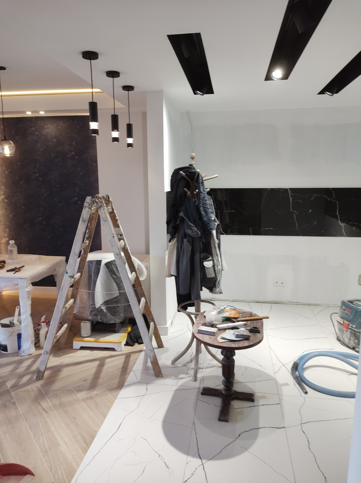 Wnętrze mieszkania w trakcie prac wykończeniowych: widoczna drabina malarska, narzędzia na stoliku, częściowo wykończona ściana z czarnymi płytami imitującymi marmur oraz lampy sufitowe.