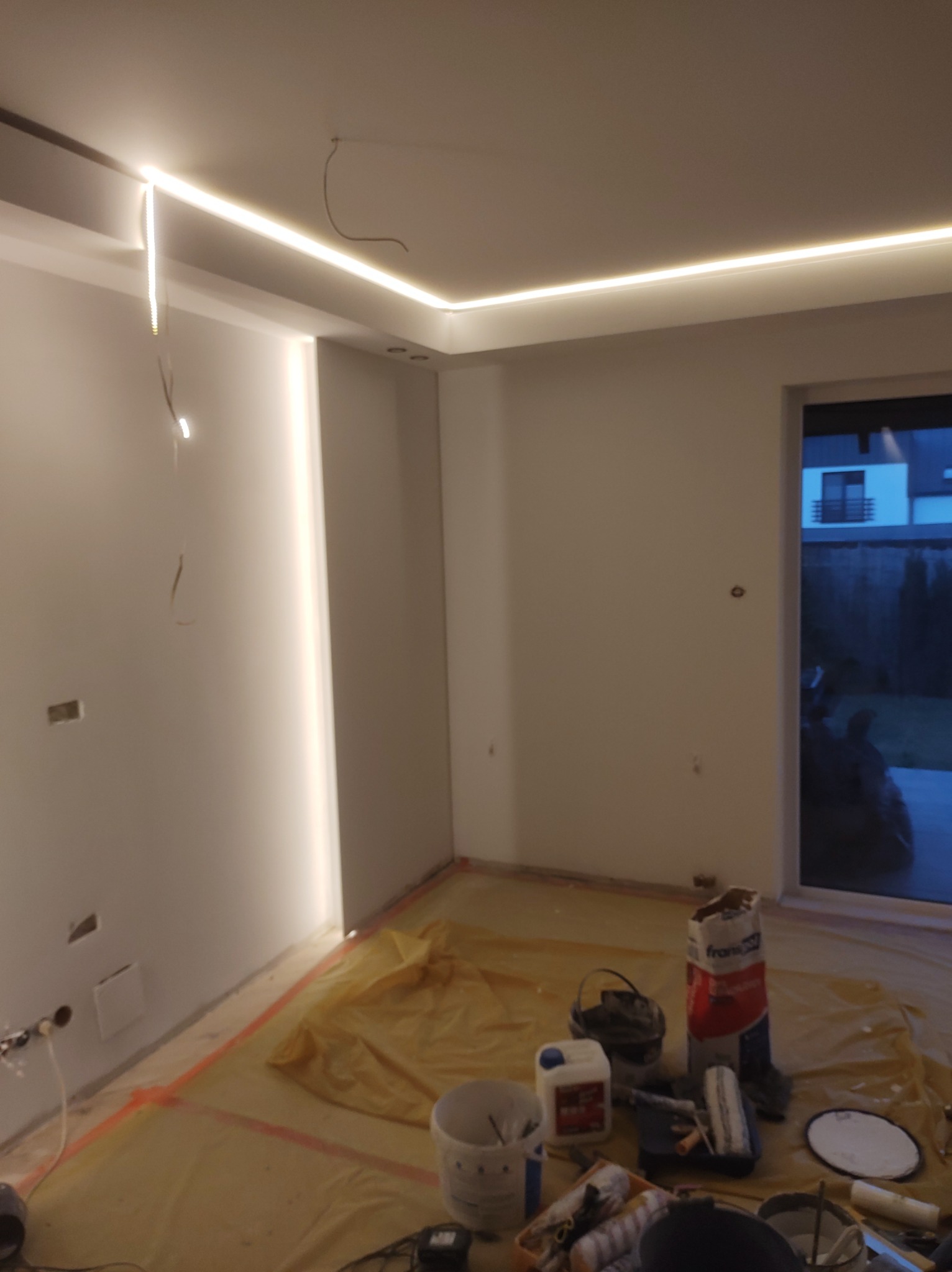 Pokój w trakcie prac wykończeniowych, z zamontowanym oświetleniem LED w suficie podwieszanym, zabezpieczony folią malarską, widoczne narzędzia i materiały malarskie.