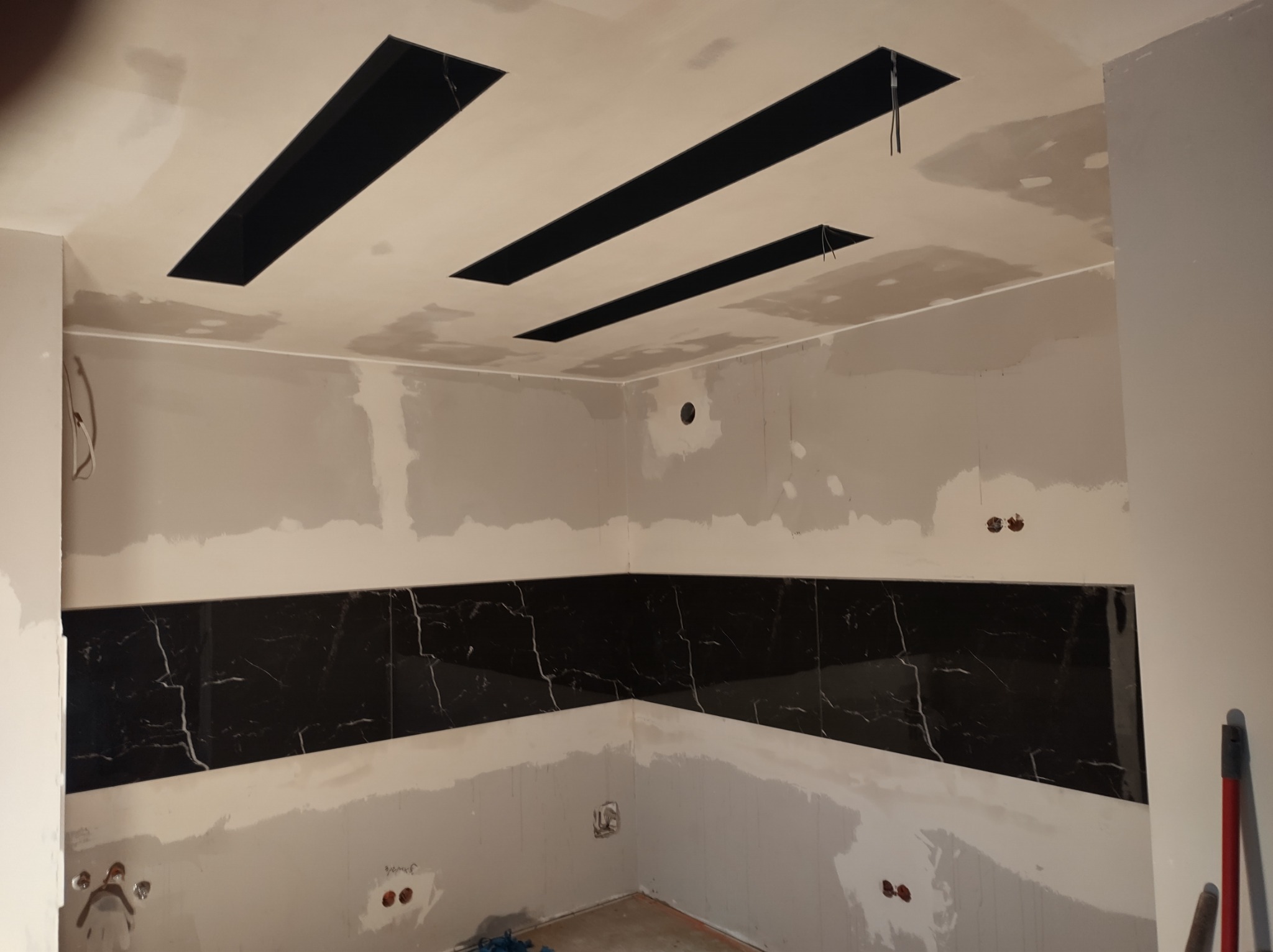 Wnętrze w trakcie prac wykończeniowych: czarne, marmurowe płytki ułożone w pasie na ścianie, sufit z otworami na oświetlenie, ściany przygotowane do malowania, widoczne ślady szpachlowania.