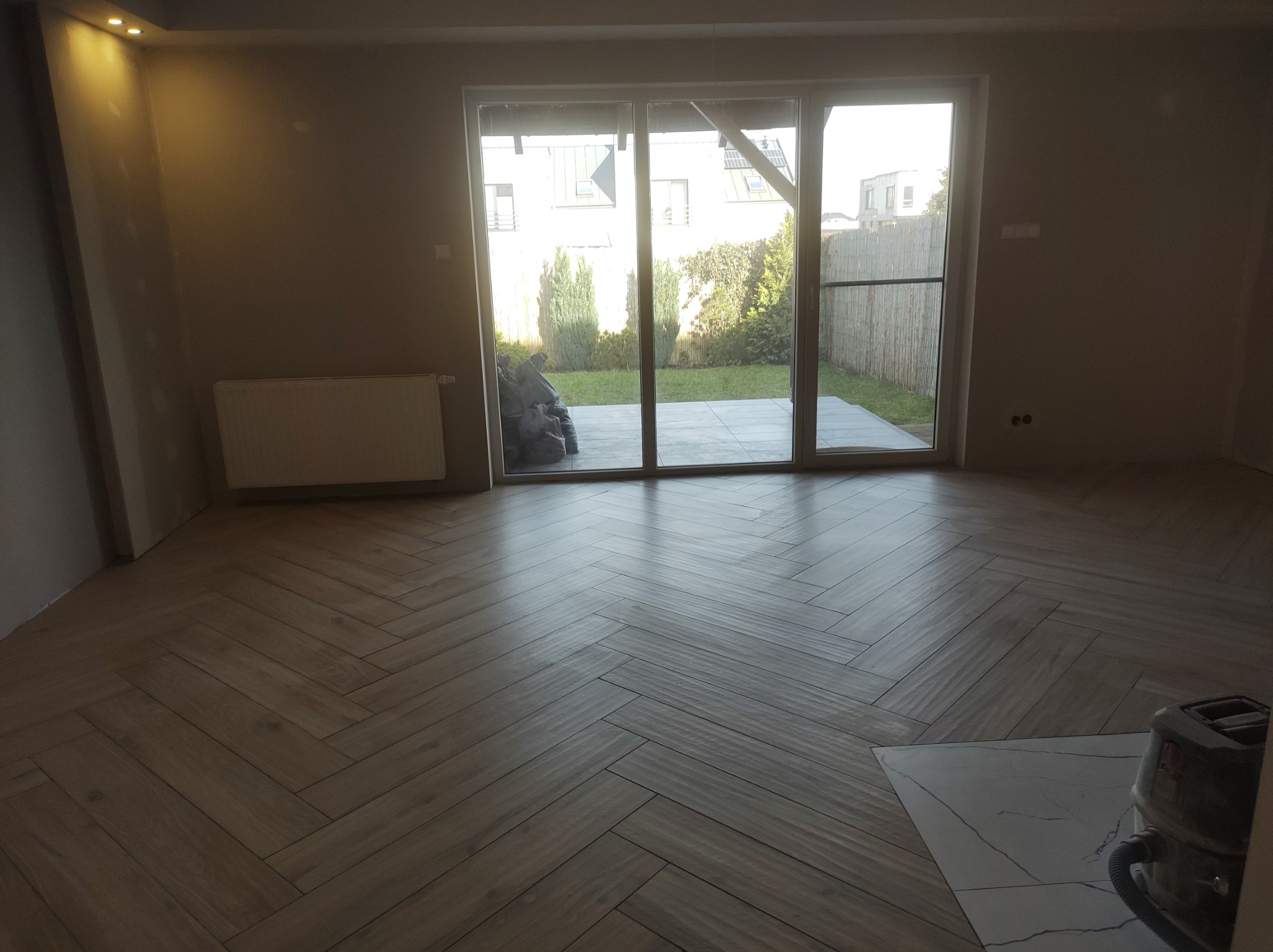 Ułożona podłoga z paneli w jodełkę w nowym pokoju z dużym oknem tarasowym wychodzącym na ogród, widoczny grzejnik na ścianie i odkurzacz przemysłowy.