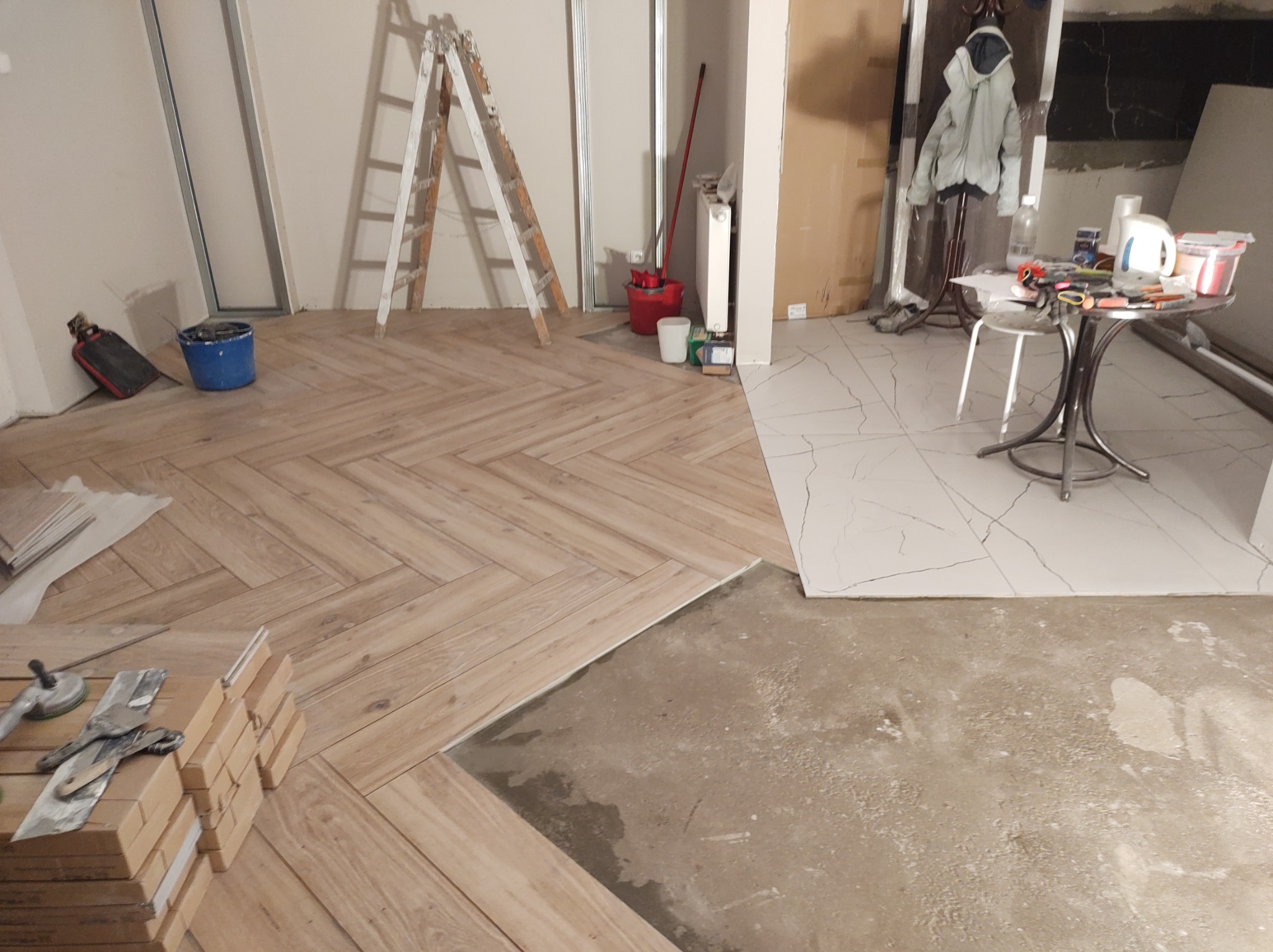 Układanie drewnopodobnych płytek w jodełkę, widoczne narzędzia, resztki kleju i fragment podłogi z białych płytek imitujących marmur.