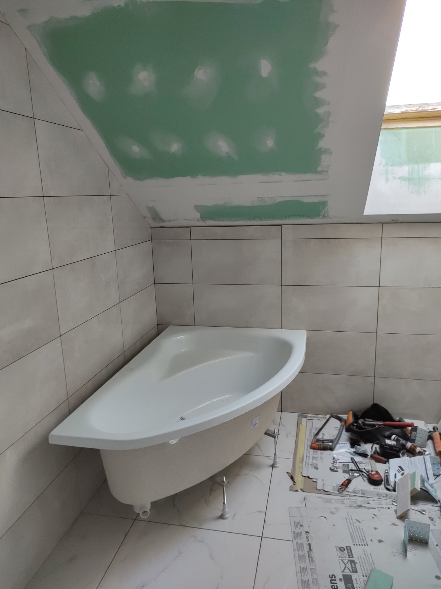 Narożna wanna na nóżkach w trakcie montażu w łazience z szarymi płytkami, sufit z płyt karton gips, widoczne narzędzia i materiały budowlane.