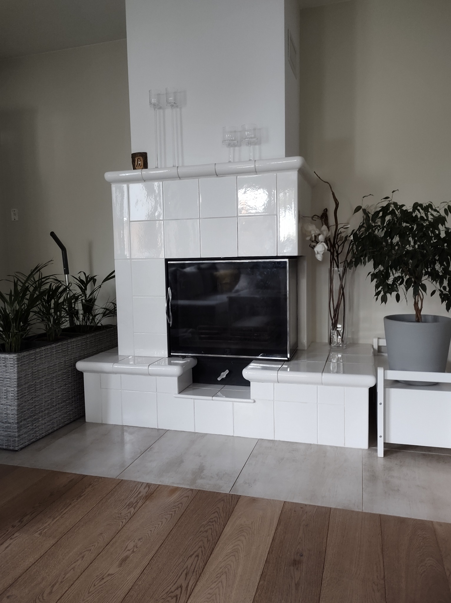 Biały, kaflowy kominek w jasnym wnętrzu, z ozdobnymi świecznikami, roślinami w donicach i drewnianą podłogą.