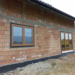 Montaż okien z brązowymi ramami w surowym murze z cegły ceramicznej, widoczne zabezpieczenie fundamentów folią kubełkową.