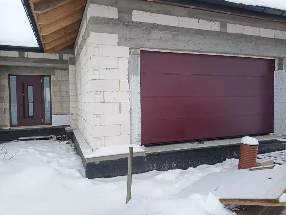 Czerwona brama segmentowa garażowa w surowym stanie budynku z białego bloczka, zasypanym śniegiem.