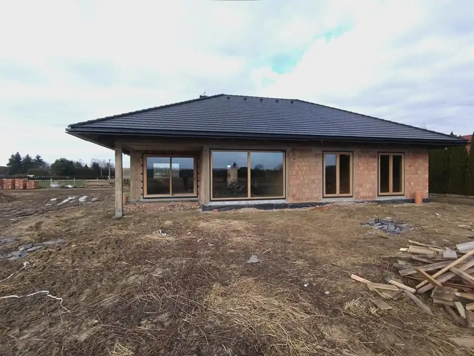 Surowy stan domu jednorodzinnego z zamontowanymi oknami PCV o drewnopodobnej ramie, widoczne cegły i przygotowany teren.
