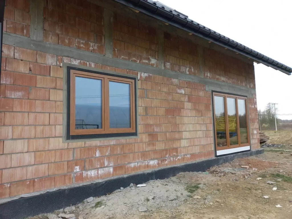 Montaż okien z brązowymi ramami w surowym murze z cegły ceramicznej, widoczne zabezpieczenie fundamentów folią kubełkową.