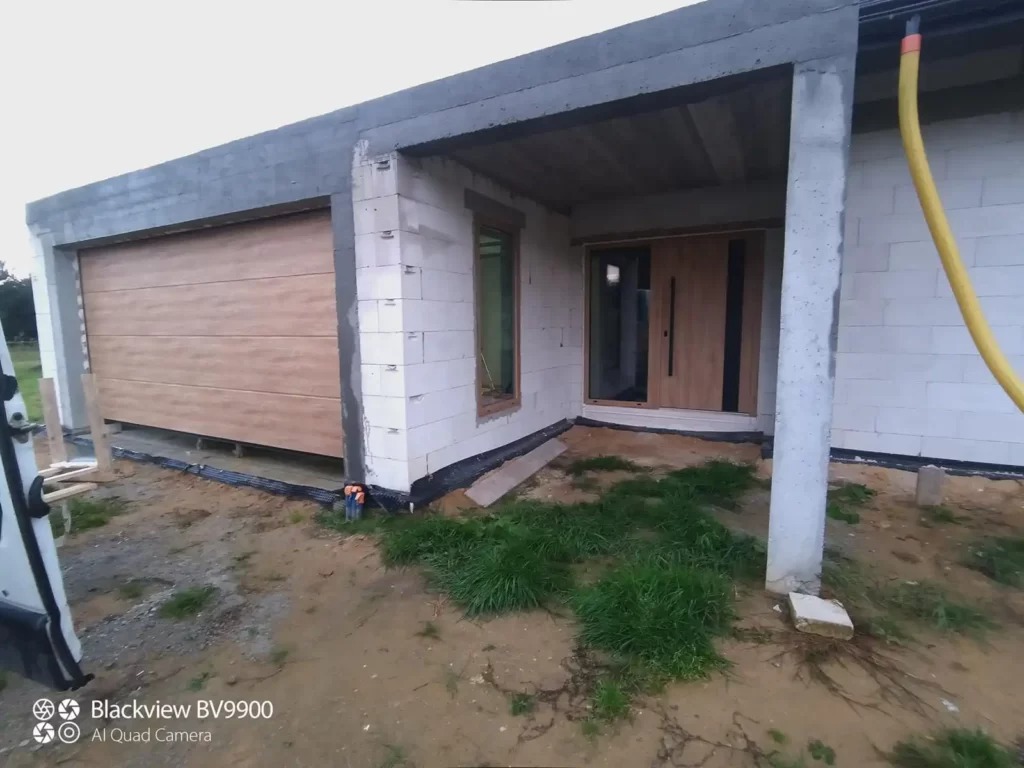 Montaż jasnej, segmentowej bramy garażowej z poziomym wzorem drewna w surowym stanie budynku z pustaków, widoczna instalacja rynny i drzwi wejściowe obok.