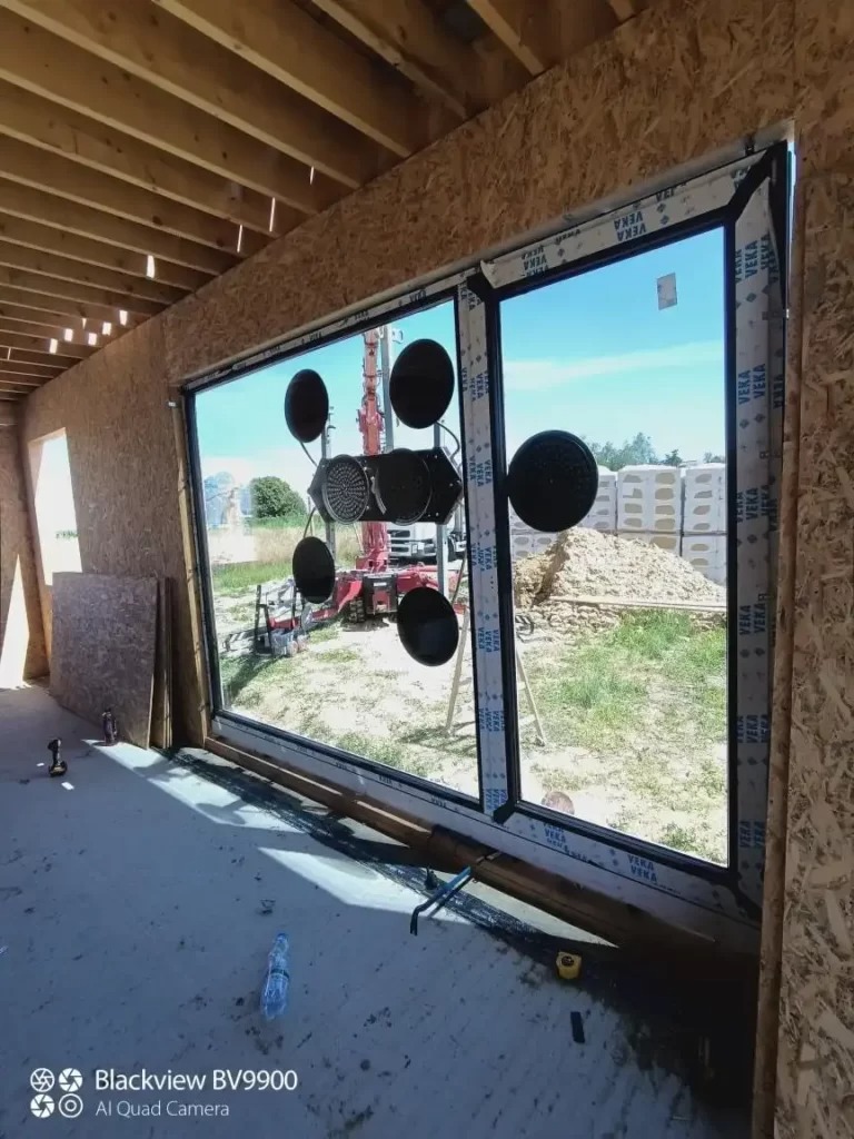 Montaż dużego okna z czarną ramą za pomocą przyssawek w surowym stanie budynku, widoczny dźwig na zewnątrz.