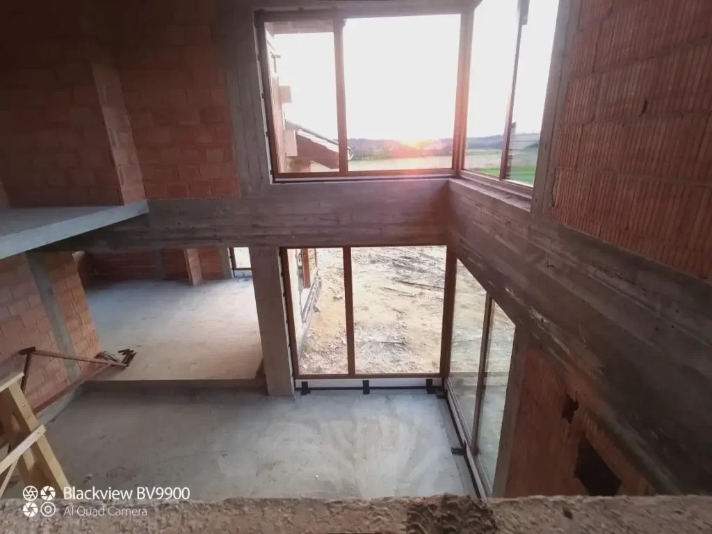 Surowy stan budynku w trakcie budowy, duże przeszklenia okienne z widokiem na zachodzące słońce, ściany z czerwonej cegły, betonowe nadproża, drabina w rogu pomieszczenia.