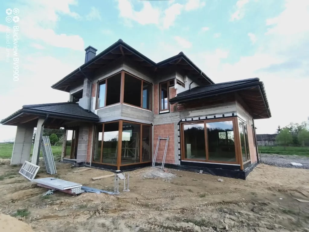 Surowy stan domu jednorodzinnego z zamontowanymi oknami z drewnopodobną ramą, widoczne rusztowania i materiały budowlane na placu budowy.