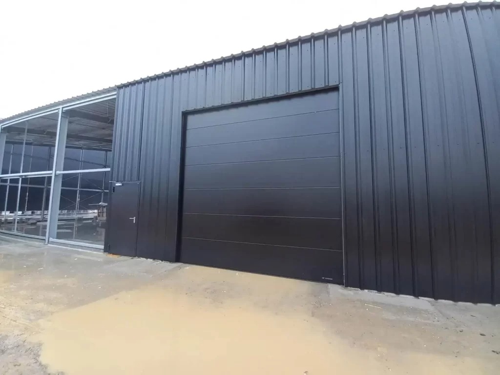 Ciemnoszara segmentowa brama garażowa w metalowej hali, z widocznym fragmentem szkieletu innej części budynku i kałużami na zewnątrz.