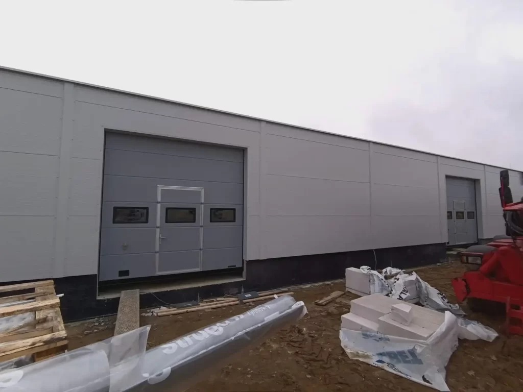 Szara brama segmentowa z przeszkleniami w hali przemysłowej, widoczne materiały budowlane i fragment czerwonego pojazdu.