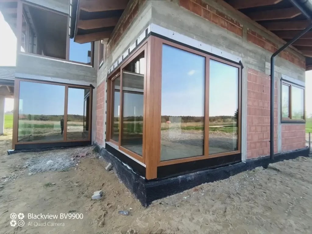 Budynek w trakcie budowy z zamontowanymi oknami z brązowymi ramami, widoczne cegły i piasek na zewnątrz.