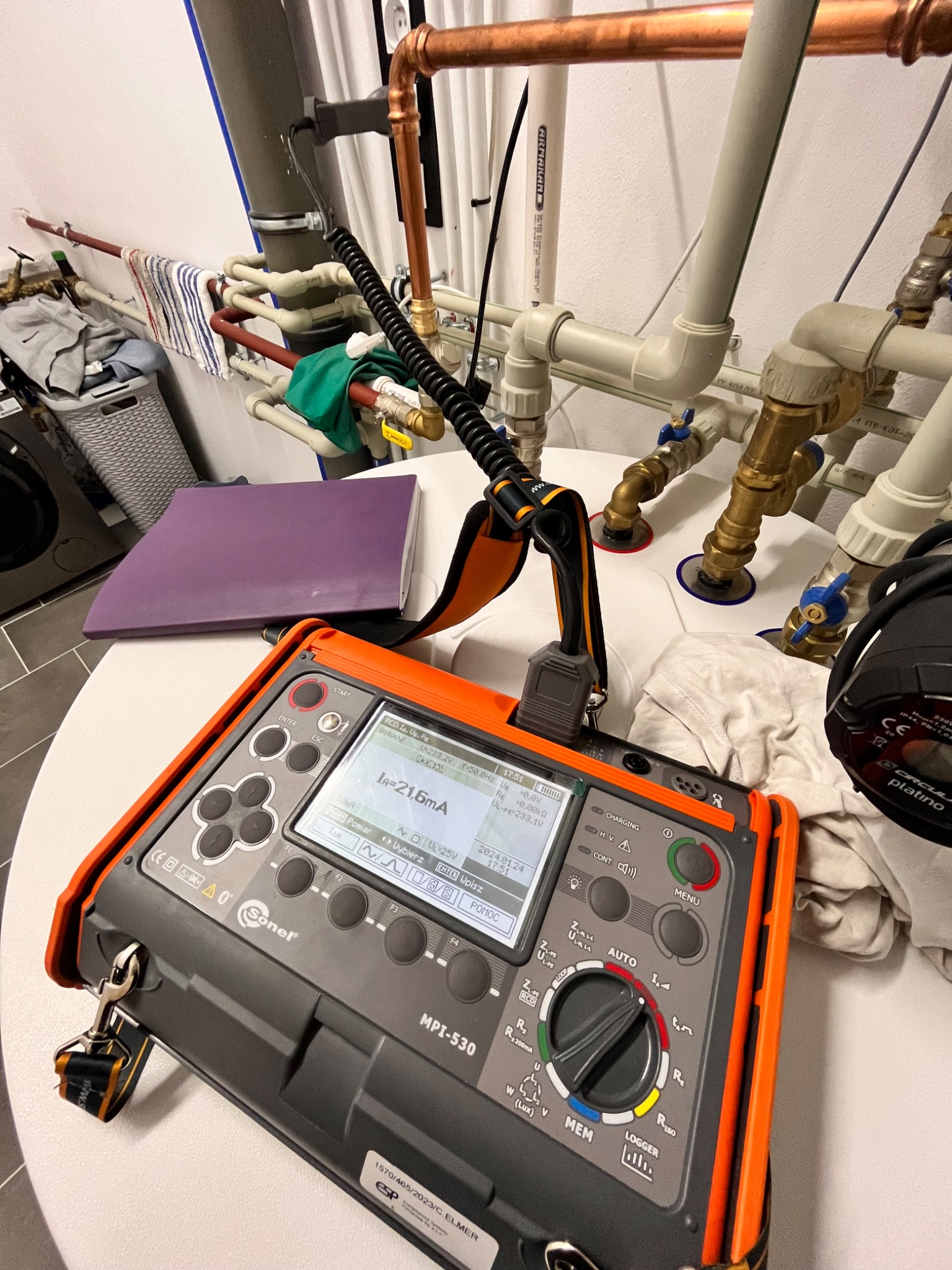 Miernik Sonel MPI-530 na białym stole, widoczne rury instalacji wodnej w tle, fioletowy segregator i pralka.