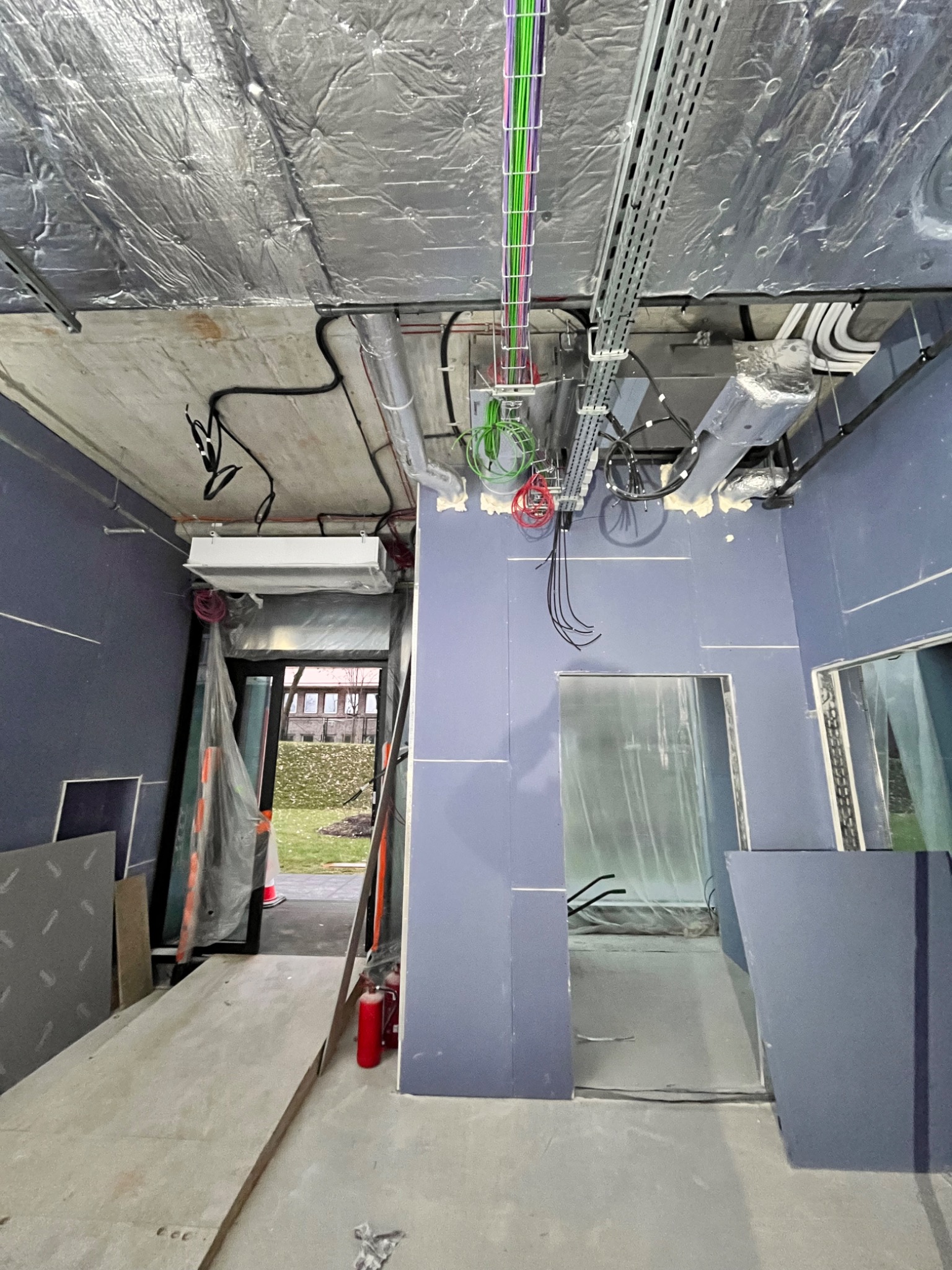 Wnętrze w trakcie wykańczania z widocznymi przewodami elektrycznymi w korytach pod sufitem, instalacją wentylacyjną i klimatyzacją, ściany w kolorze lila, podłoga betonowa, w tle widoczny trawnik...