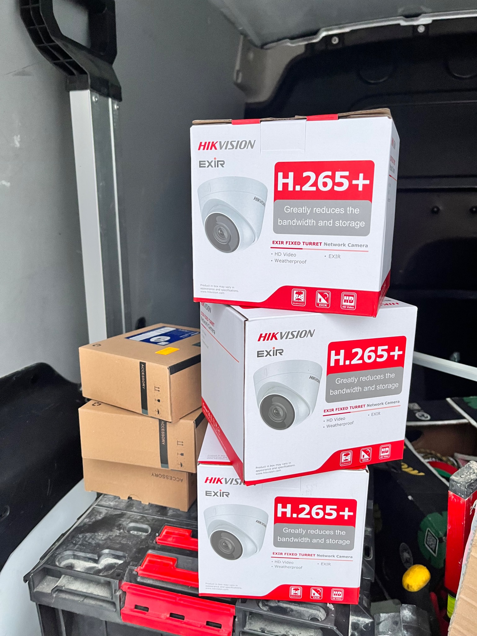 Stos pudełek z kamerami sieciowymi Hikvision EXIR Fixed Turret H.265+ w bagażniku samochodu dostawczego, obok akcesoriów w kartonach i narzędzi.