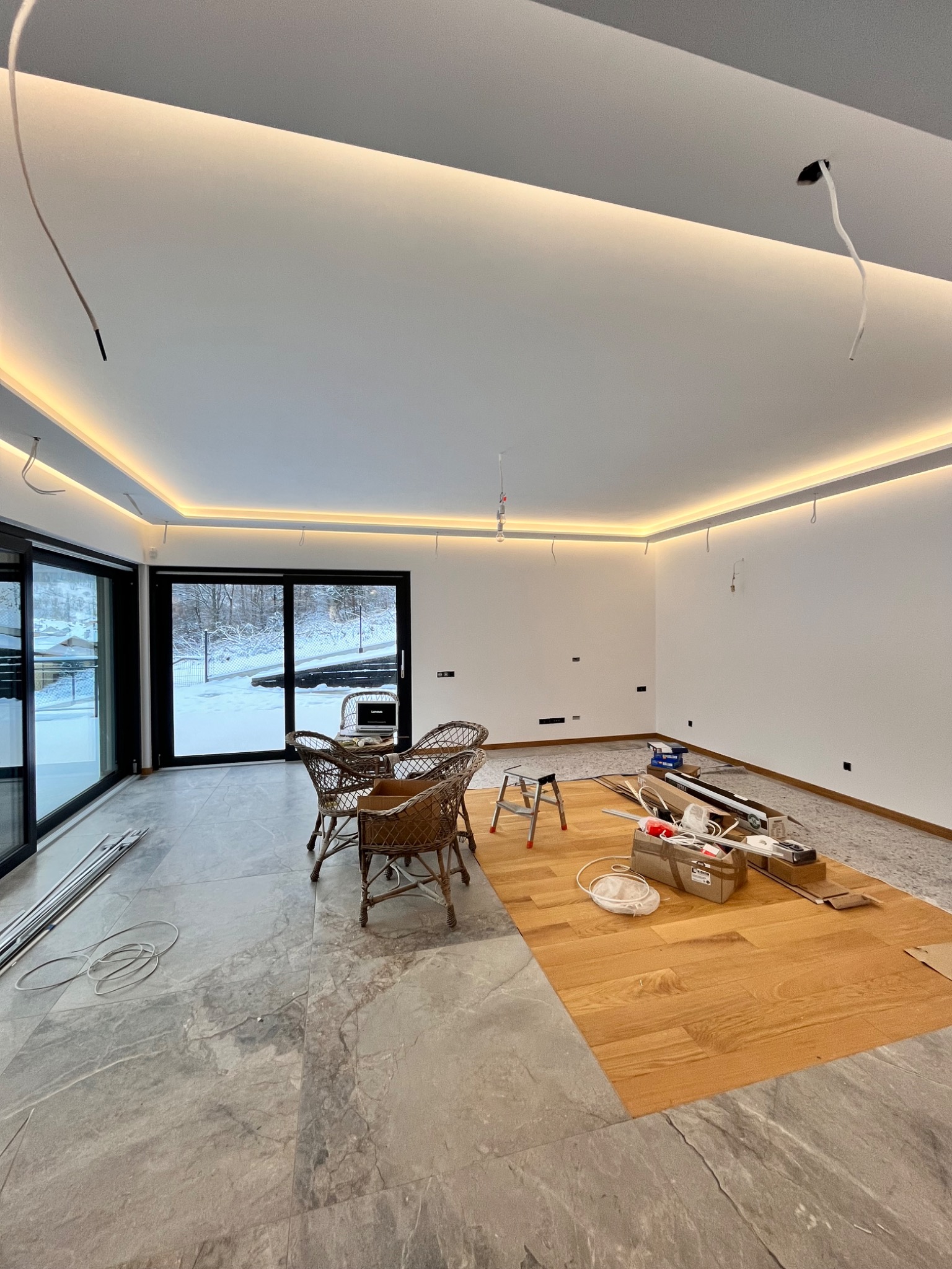 Wnętrze w trakcie wykańczania z widocznymi kablami wystającymi z sufitu pod oświetleniem LED, narzędzia pracy na podłodze, częściowo ułożona podłoga drewniana oraz widok na zaśnieżony ogród przez...