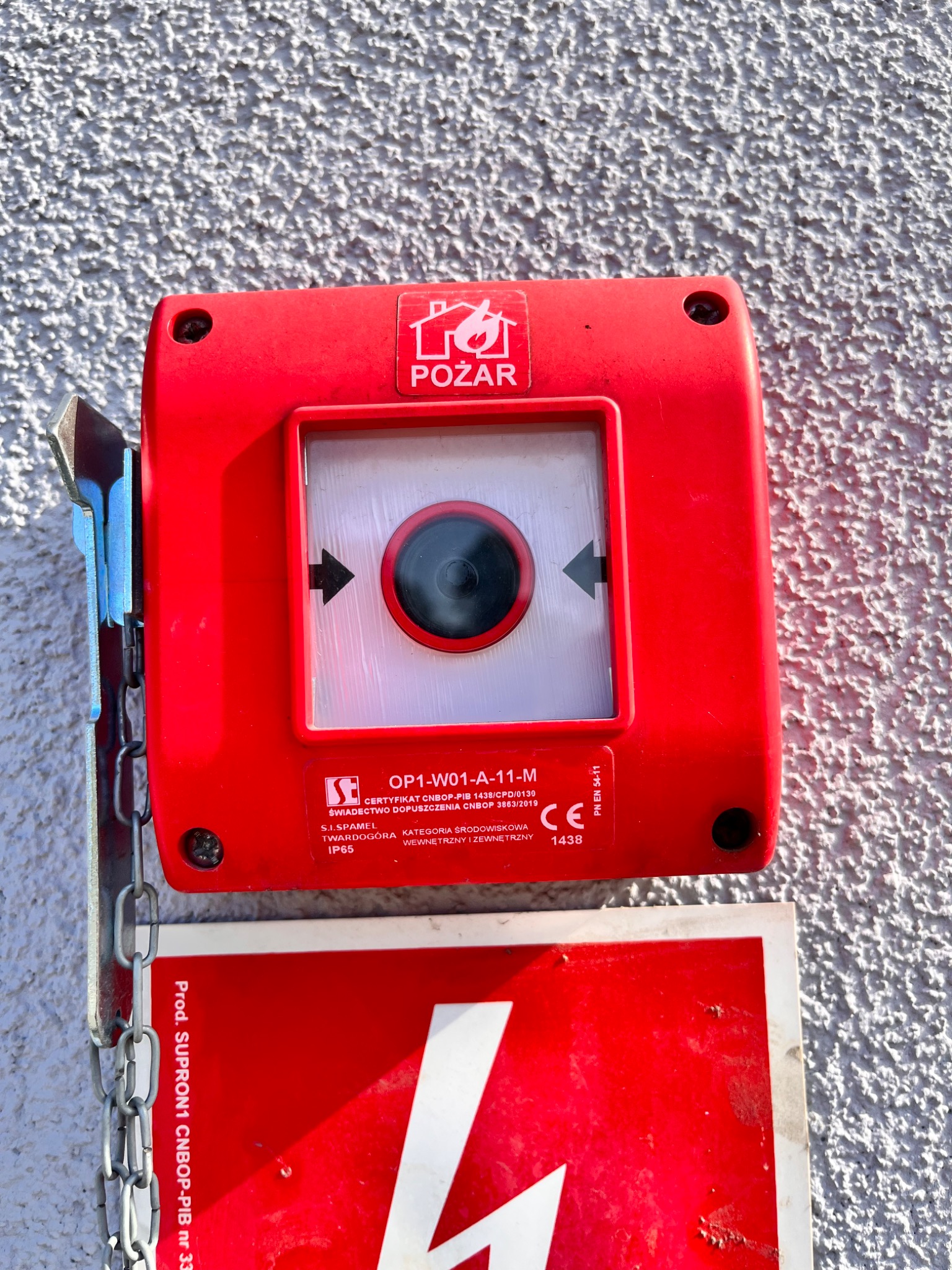 Czerwony, natynkowy ręczny ostrzegacz pożarowy OP1-W01-A-11-M z logo 'Pożar', zamontowany na szarej, chropowatej ścianie, z widoczną tabliczką informacyjną poniżej i metalowym uchwytem z łańcuchem...