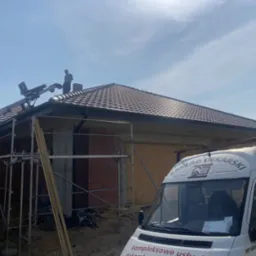 Dekarz na dachu domu jednorodzinnego z ciemną dachówką, widoczna podnośnik do dachówki, rusztowanie i samochód firmowy.