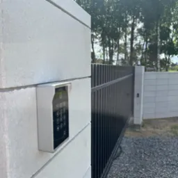 Zewnętrzny panel numeryczny do otwierania bramy przesuwnej, zamontowany na słupku z szarego betonu komórkowego, widok na czarną bramę i otoczenie z drzewami w tle.