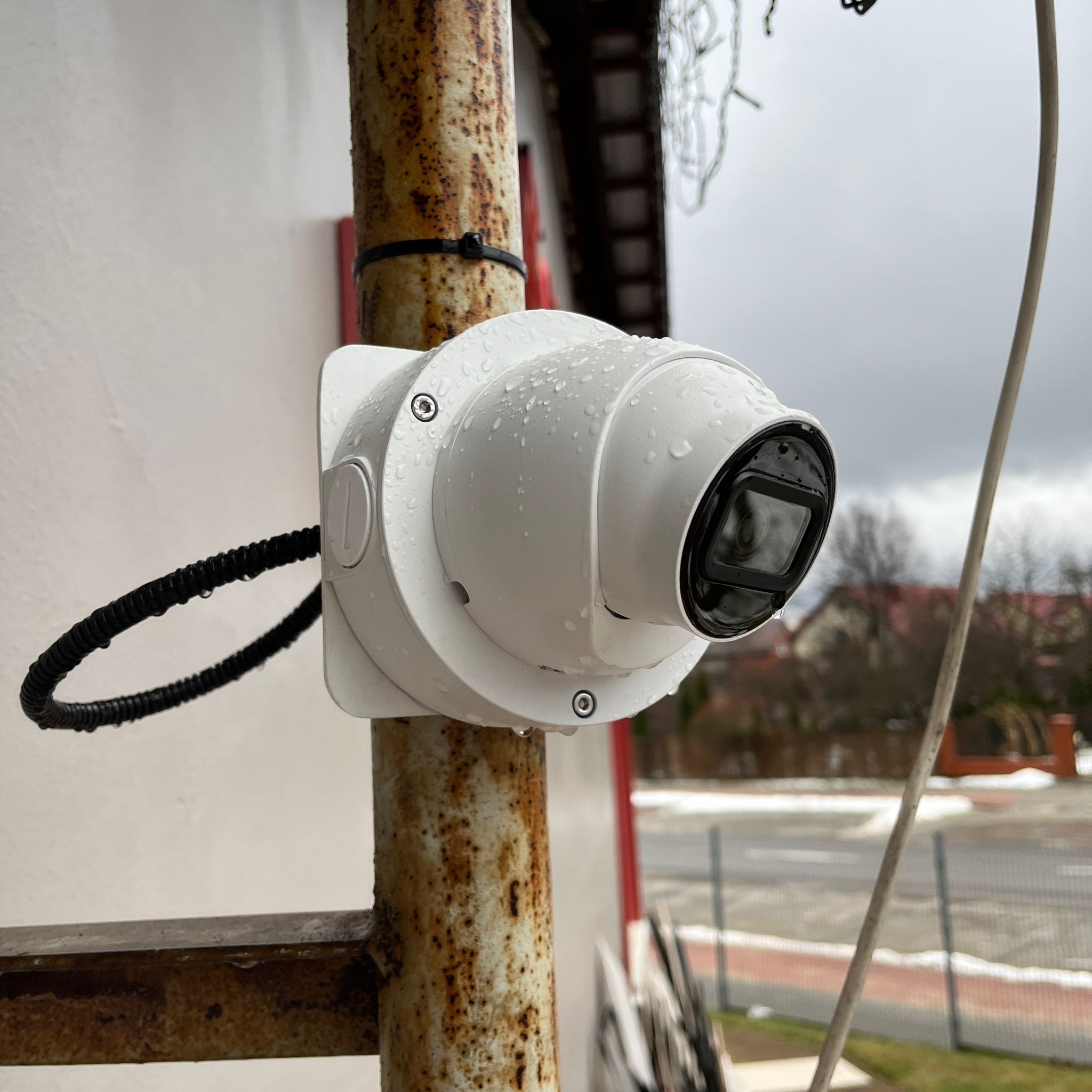 Biała kamera monitoringu PTZ z kroplami deszczu, zamontowana na zardzewiałej metalowej rurze na zewnątrz budynku.