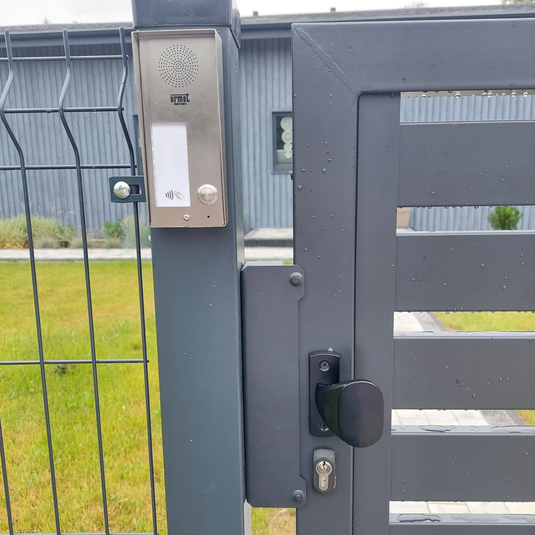 Szara furtka z panelem domofonu Urmet z czytnikiem RFID, widoczne krople wody po deszczu, w tle ogrodzenie panelowe i fragment domu.