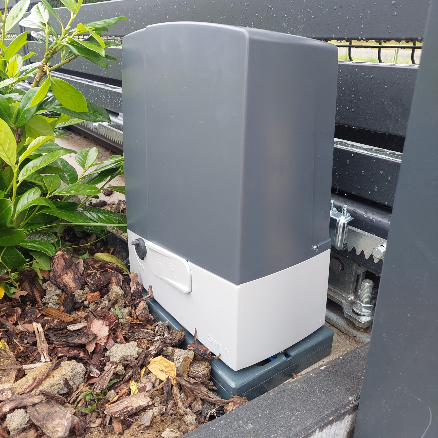 Szary automat do bram przesuwnych zamontowany w Opocznie, widoczny mechanizm zębatkowy i fragment ogrodzenia, obok roślinność.