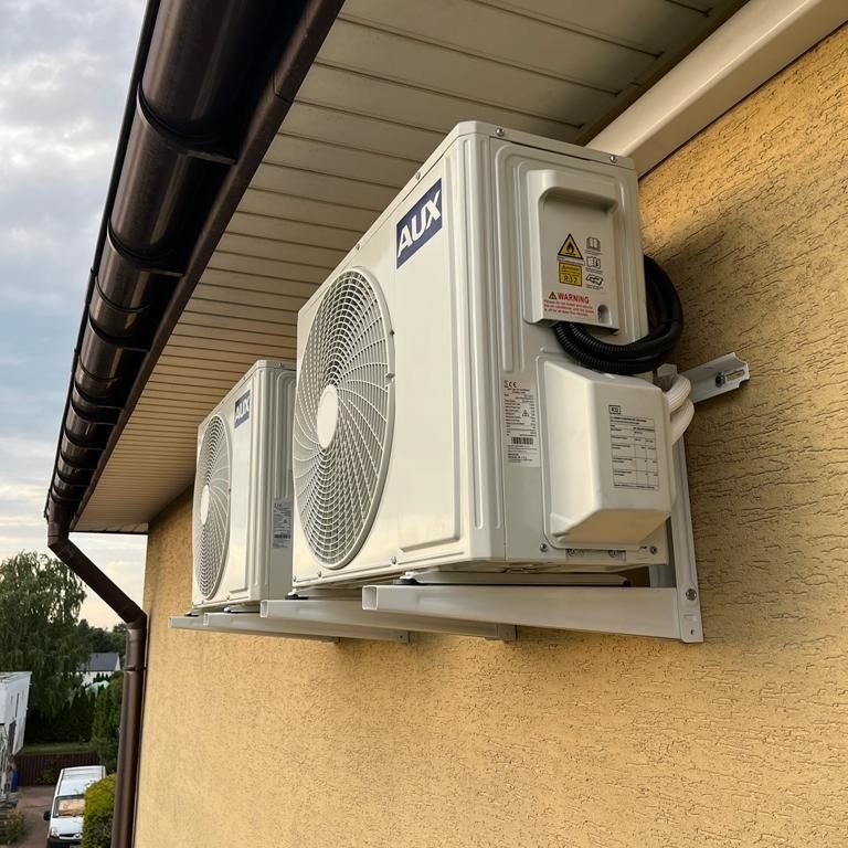 Dwa białe klimatyzatory marki AUX zamontowane na elewacji domu z brązowym dachem, widoczne kable i uchwyty montażowe.