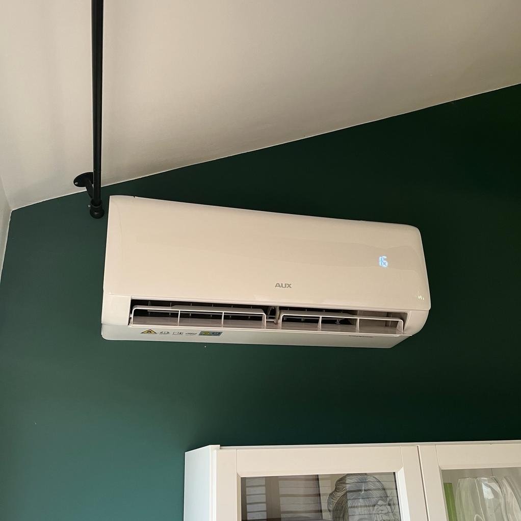 Biały klimatyzator marki AUX zamontowany na zielonej ścianie nad białymi szafkami. Na wyświetlaczu klimatyzatora widoczna temperatura 16 stopni.
