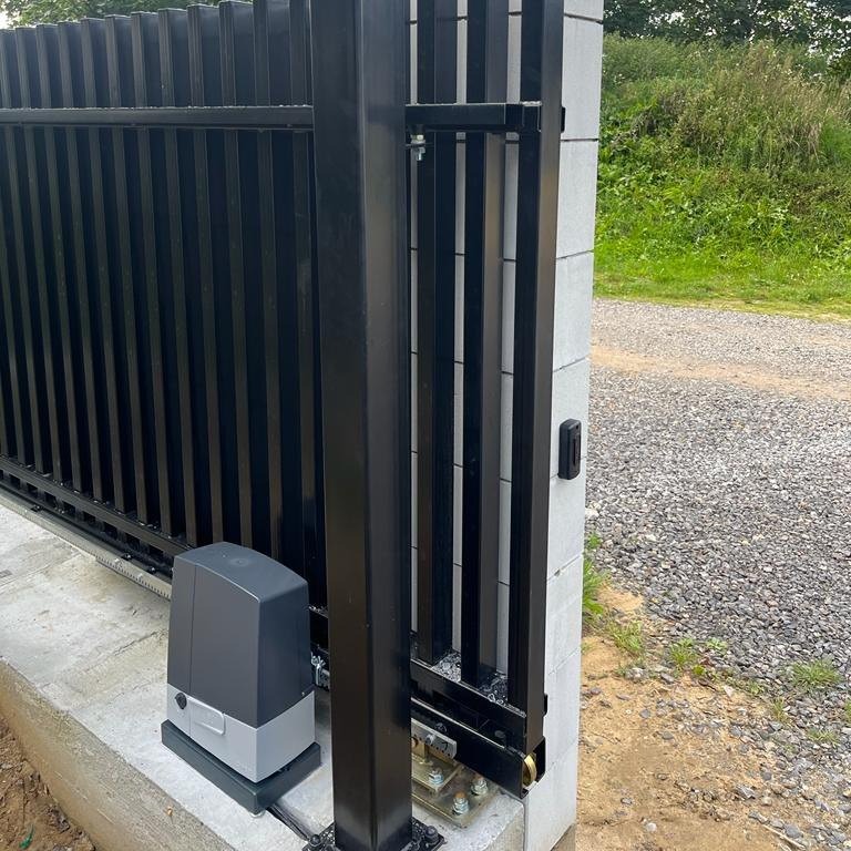 Czarna, przesuwna brama z automatyką, widoczny napęd bramy w szarej obudowie zamontowany na betonowej podstawie, szyny prowadzące bramę, tło stanowi żwirowa droga i zieleń.