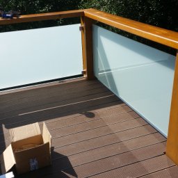 FRB JANICKI - Balkon z brązową deską kompozytową i szklaną balustradą. Widoczny karton i narzędzia. Tło z zielenią. Jasne oświetlenie słoneczne.