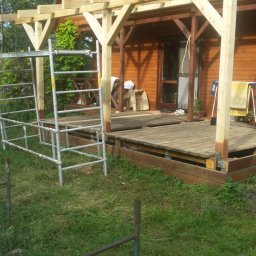 FRB JANICKI - Drewniana konstrukcja z podestem przy domu z bali, widoczne rusztowanie i trawnik. Częściowo zadaszona weranda w trakcie budowy.