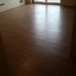 Ukończone układanie paneli podłogowych w nowym mieszkaniu, widoczne gniazdka elektryczne i okno balkonowe. Brązowe panele ułożone r&oacute;wno, ściany przygotowane do malowania.