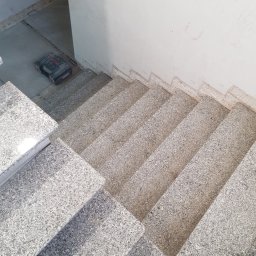 FRB JANICKI - Wykończone granitowe schody wewnętrzne z widocznymi śladami po pomiarach na ścianie, skrzynka narzędziowa w tle. Widok z góry, fragmentarycznie widoczne buty.