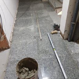 FRB JANICKI - Układanie szarej, nakrapianej terakoty w korytarzu. Widoczne narzędzia: poziomica, laser krzyżowy, wiadro z zaprawą. Etap prac: częściowo ułożone płytki, pozostałe do ułożenia.