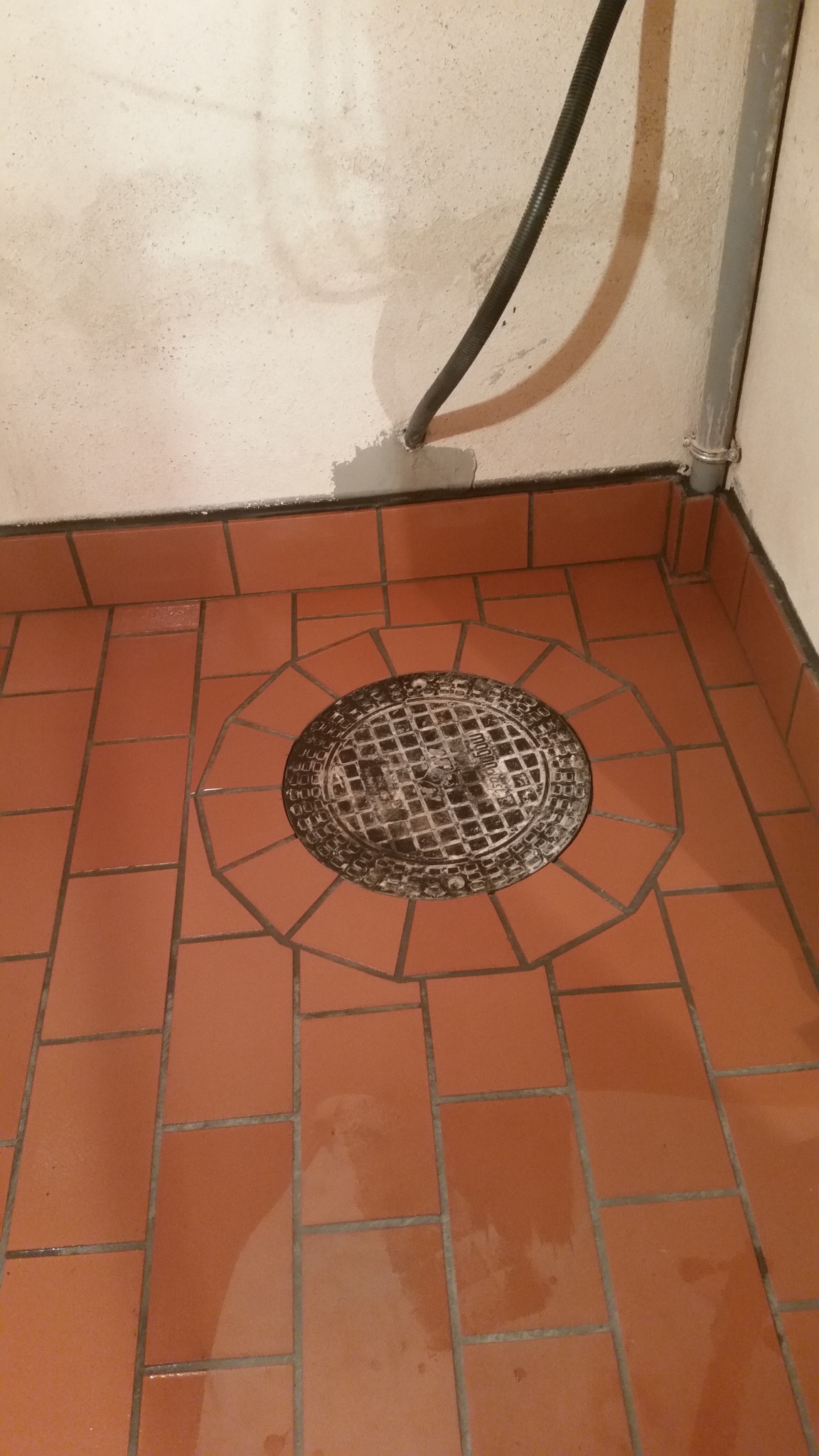 Pomarańczowa podłoga z terakoty z okrągłym odpływem kanalizacyjnym pośrodku, widoczne rury przy ścianie.