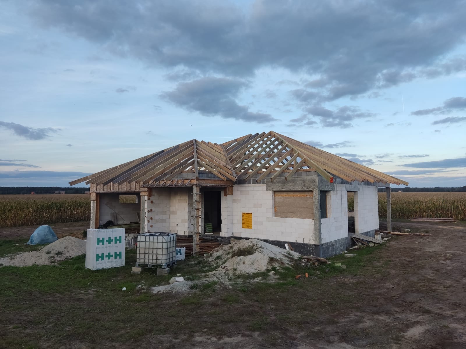 Budowa domu jednorodzinnego: surowy stan otwarty z widoczną konstrukcją dachu, bloczki H+H i piasek na działce, pole w tle, pochmurne niebo.