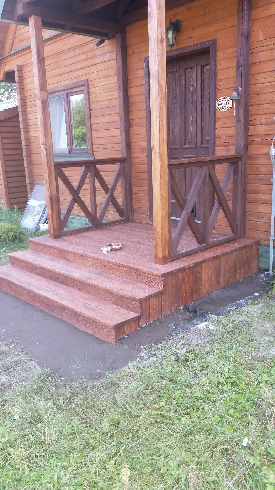 Drewniany ganek z balustradą i trzema stopniami, świeżo po impregnacji. Widoczne detale drewnianej konstrukcji i otoczenie z zielenią.