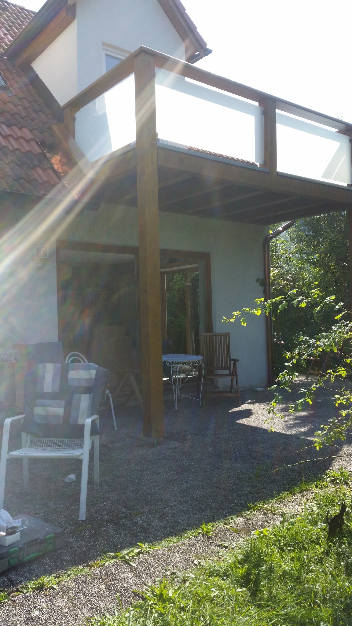 Zadaszony taras z drewnianą konstrukcją i szklanymi balustradami, widok z zewnątrz w słoneczny dzień. Widoczne meble ogrodowe i roślinność.