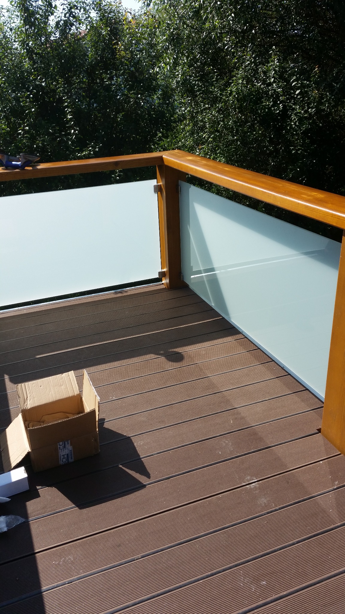 Balkon z brązową deską kompozytową i szklaną balustradą. Widoczny karton i narzędzia. Tło z zielenią. Jasne oświetlenie słoneczne.