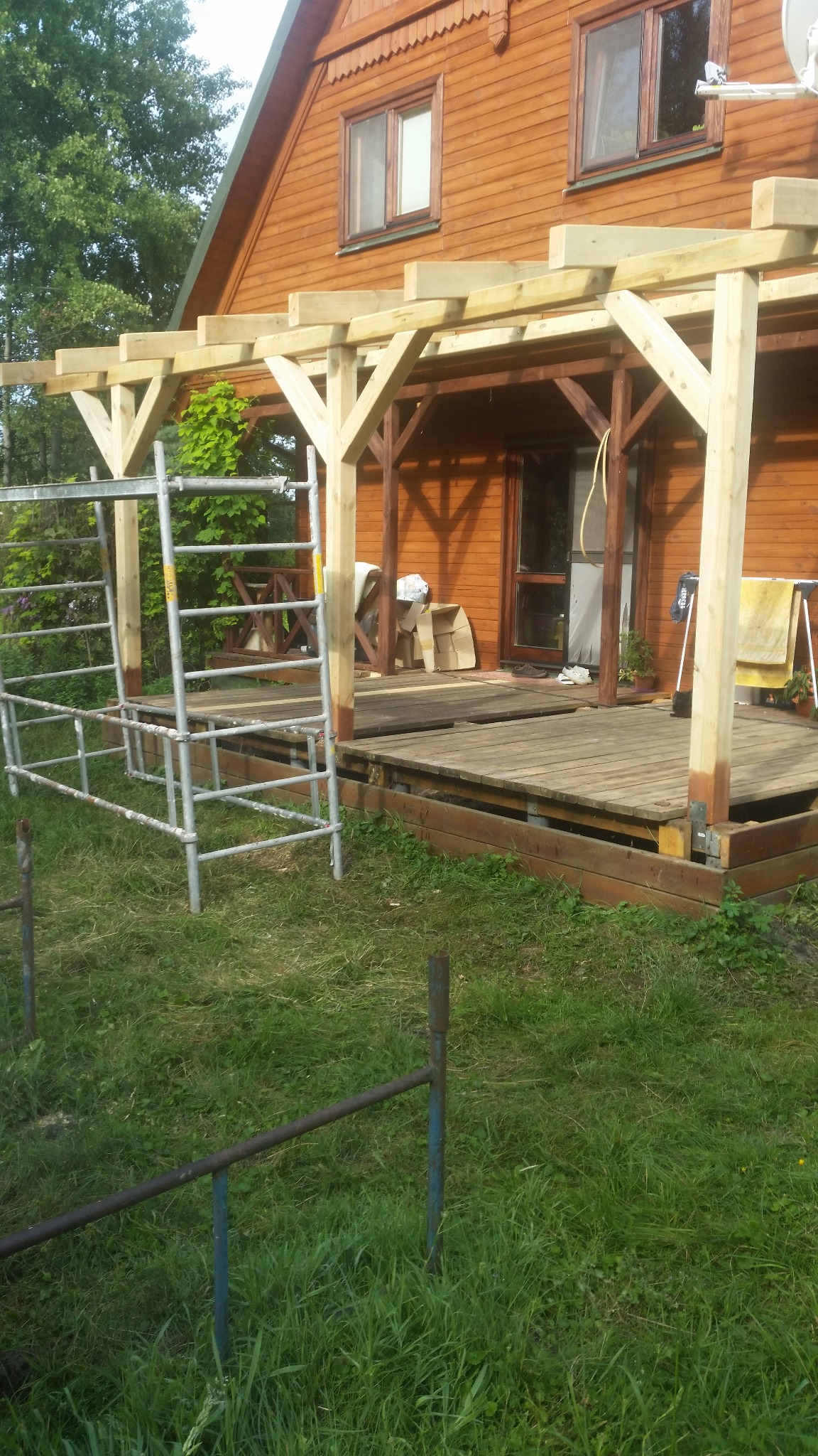 Drewniana konstrukcja z podestem przy domu z bali, widoczne rusztowanie i trawnik. Częściowo zadaszona weranda w trakcie budowy.